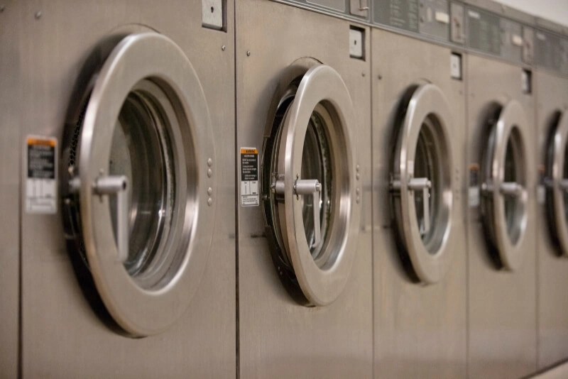 Row of industrial washing machines with closed round doors, lined up in a laundromat, displaying laundry pricing and a shallow depth of field, Laundry Services in Toronto