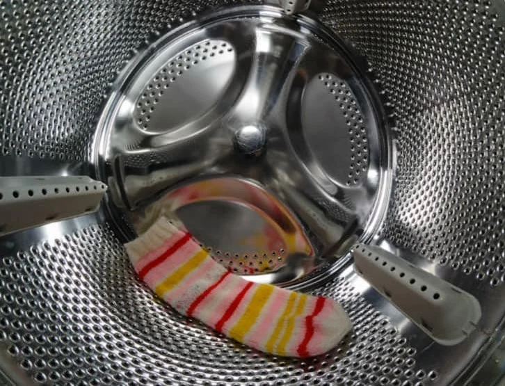 A single striped sock lies inside an industrial stainless steel washing machine drum with holes and a visible central agitator.