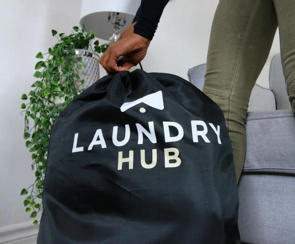 Person holding a black laundry bag labeled "Laundry Hub" in a room with a gray sofa and a green plant.