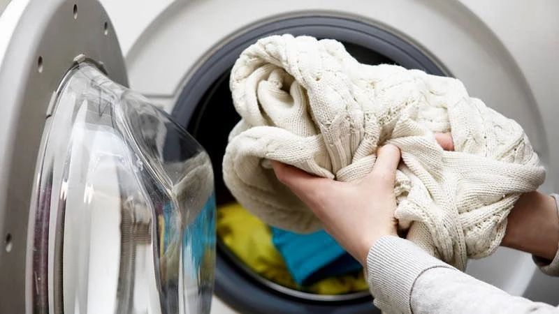Hands placing a white blanket into a front-loading washing machine in Markham, with colorful clothes visible inside.