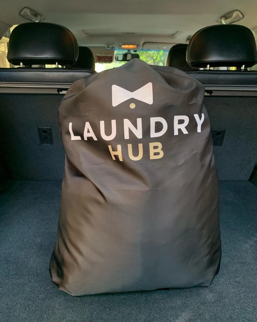A large, black laundry bag with "Laundry Pickup and Delivery in Toronto" and a white bow tie graphic, positioned in the trunk of a car.