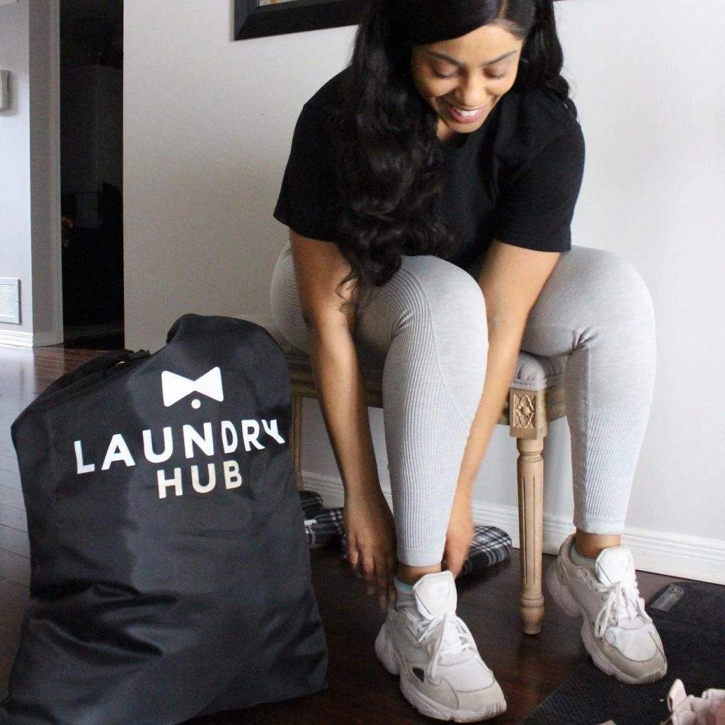 A woman in athletic wear smiles as she ties her shoelaces, sitting next to a "Laundry Pickup and Delivery in Toronto" branded bag.