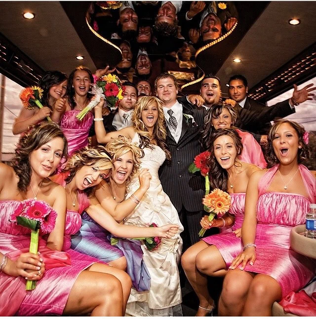 A lively wedding party in a limousine, with the bride and groom in the center surrounded by guests in cheerful poses, heading to Salon Obscur for the reception.