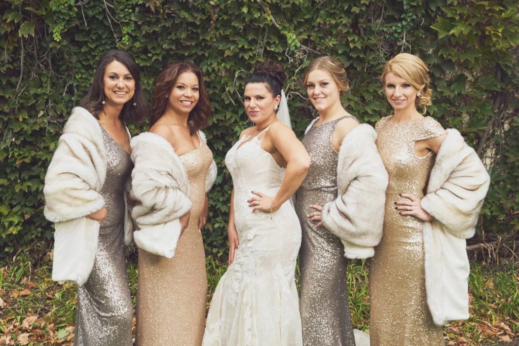 Five women in elegant dresses and faux fur coats pose outdoors near Salon Obscur, with the central figure in a white bridal gown.