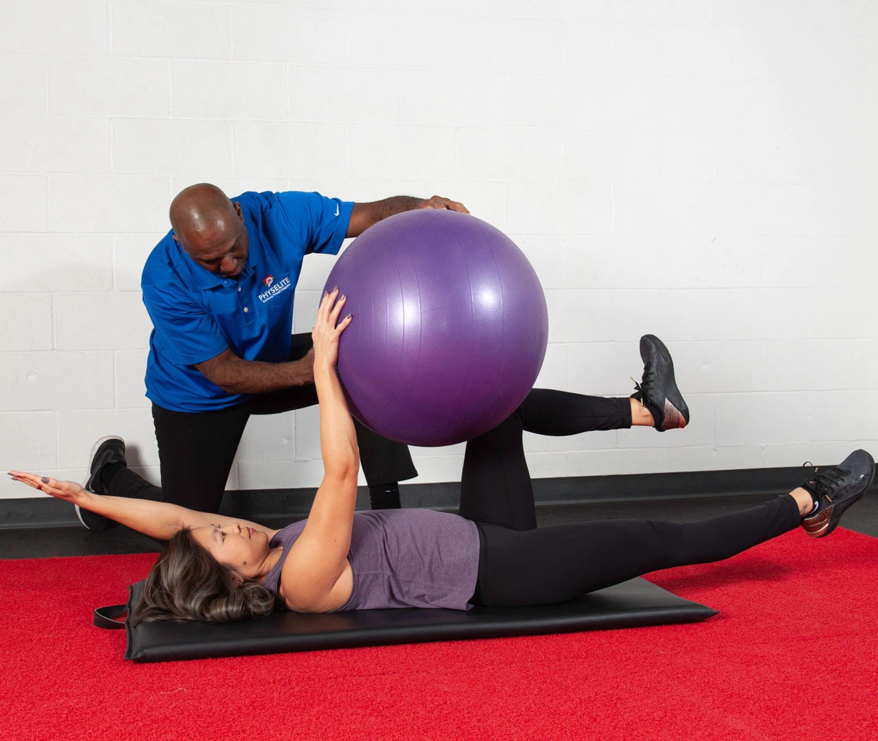 A personal trainer assists a woman performing a leg stretch with a purple exercise ball at the Laundry Hub, featuring red flooring.