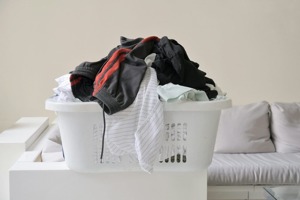 Plastic laundry basket filled with various crumpled clothes, placed on a white surface in a room with a light-colored sofa in the background, awaiting pickup by a laundry service, Contact laundry hub