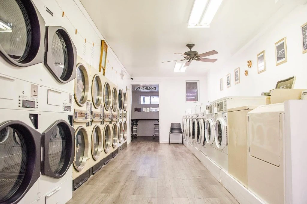 Interior of a laundromat with rows of washing machines, a tv, and a seating area under bright lighting, offering laundry pickup and delivery service.