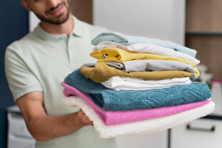 A man smiling while holding a stack of freshly folded laundry, including towels and clothes in various colors, ready for wash and fold delivery.