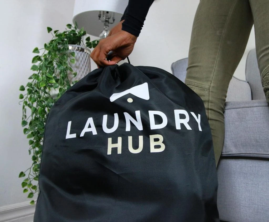 A person holding a black laundry bag labeled "Laundry Hub" in front of a gray sofa with a potted plant nearby, showcasing the convenience of Laundry Pickup and Delivery services.