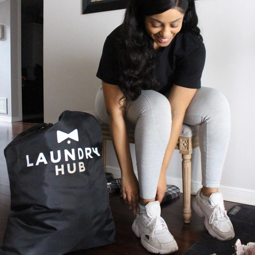 A woman sits on a chair, tying her white sneakers. Next to her is a large black laundry bag labeled "Laundry Hub," ready for Laundry Pickup and Delivery. She is wearing a black shirt and light grey leggings.