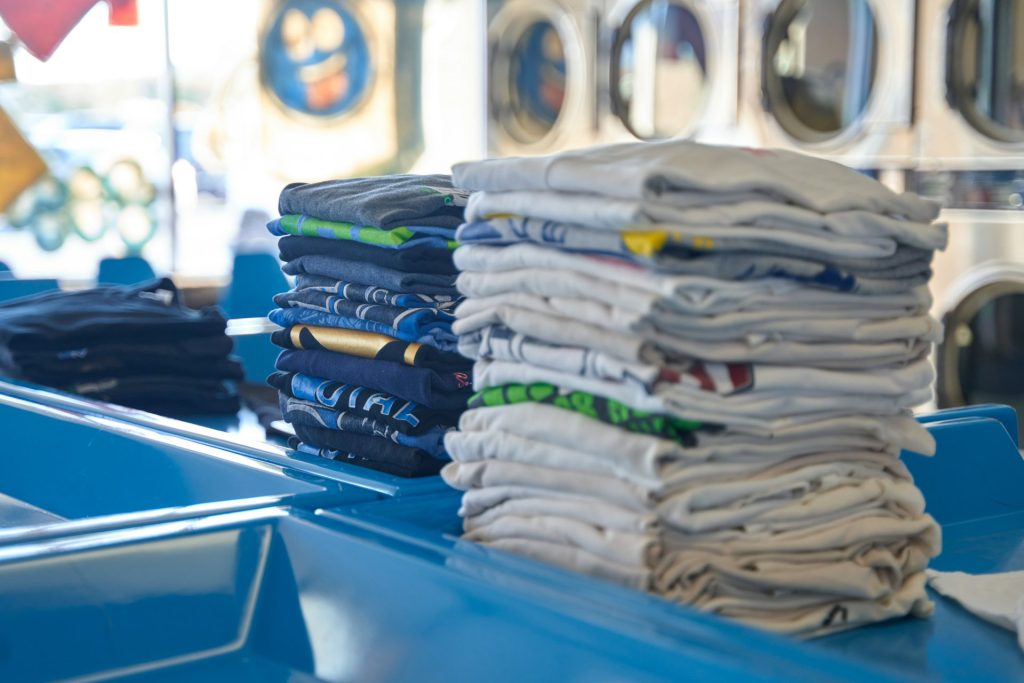 Folded clothes are stacked in multiple piles on top of blue counters in an eco-friendly laundry, with washing machines visible in the background.