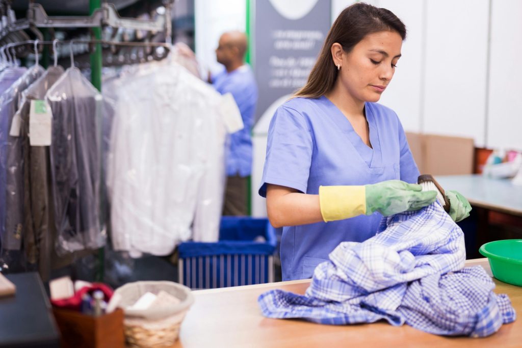 A woman in blue scrubs and yellow gloves sorts laundry in an eco-friendly dry cleaning store, with rows of hanging clothes in plastic covers in the background.