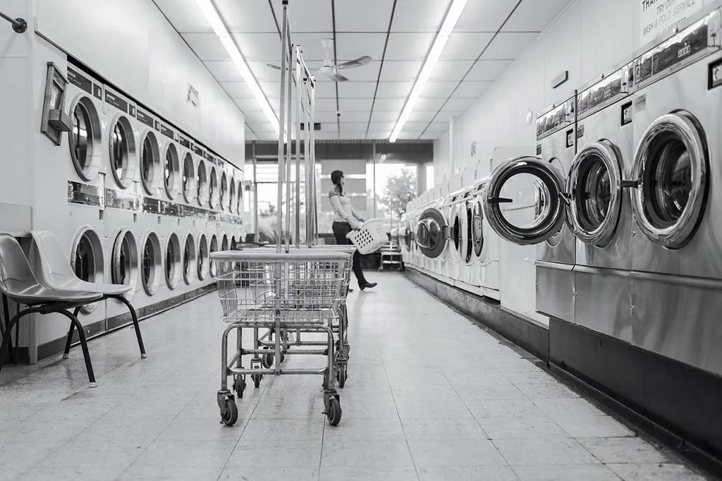 Interior of a laundromat with rows of washing machines and a shopping cart in the foreground; a person is visible in the background, utilizing the laundry solutions available.