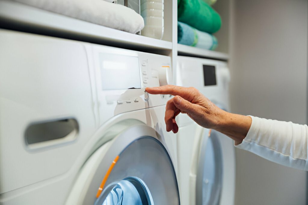 A person touches the control panel of a front-loading washing machine, with shelves of eco-friendly laundry detergent and folded towels above.