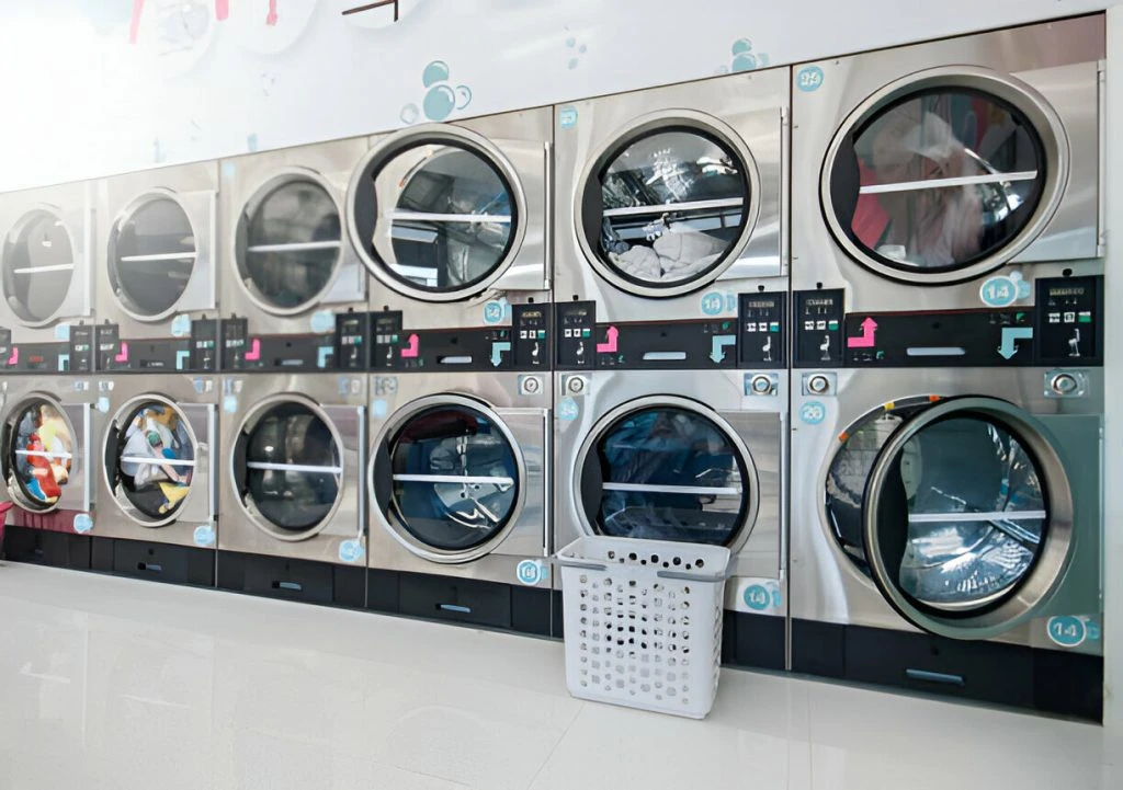 A row of ten front-loading washing machines in a laundromat, with some machines actively washing clothes and an empty laundry basket placed on the floor in front, showcasing a step towards greener dry-cleaning alternatives.