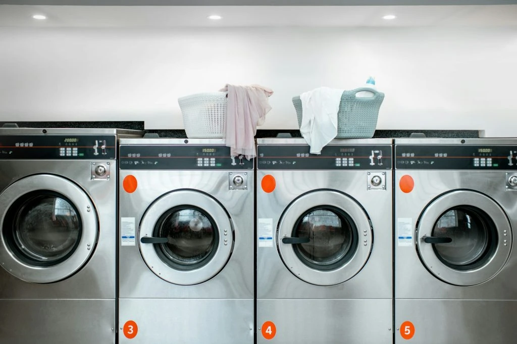 Four silver washing machines in a row, with laundry baskets placed on top. Two baskets contain clothes, and the machines are numbered 3, 4, and 5. Experience Pickering with top-quality eco-friendly laundry solutions, Pickering with top-quality eco-friendly laundry solutions