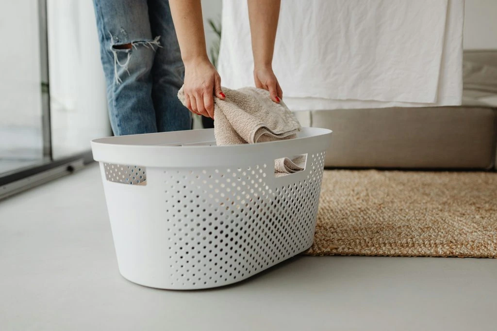 A person places a folded towel into a white plastic laundry basket, preparing it for an on-demand laundry service.