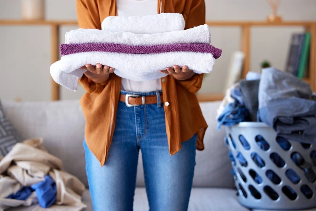 A person holding a stack of folded towels, standing in front of a couch with a laundry basket filled with clothes in the background, showcases their eco-friendly laundry tricks.