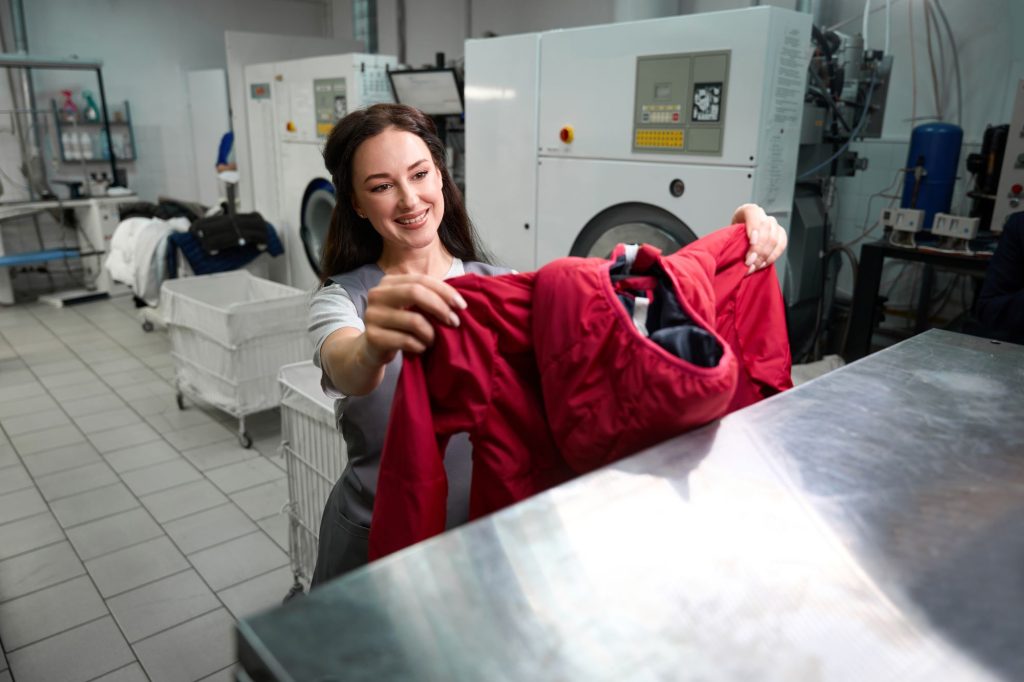 A woman inspects a red jacket in an eco-friendly laundromat in Toronto, with industrial washing machines and laundry carts visible in the background.