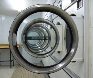 Row of industrial front-loading washing machines with open doors in a clean, white-tiled laundry room featuring wooden folding tables.