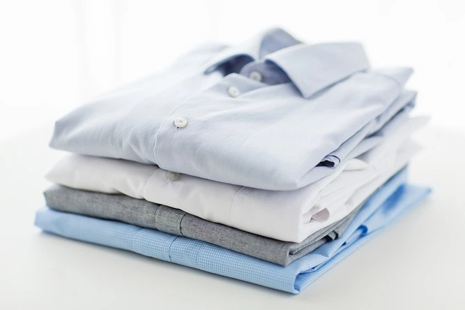 A neatly folded stack of several button-down shirts in varying shades of blue, white, and grey, each meticulously treated with sustainable dry cleaning, placed on a flat surface.