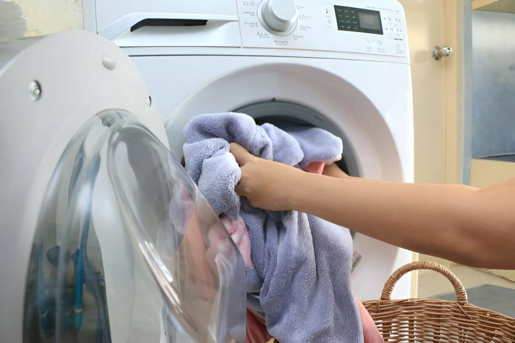 A person is loading a washing machine with laundry, placing a bundle of clothes, including a purple towel, into the open door of the front-loading washer. A wicker basket is nearby, offering an eco-friendly alternative to plastic. For delicate items, consider sustainable dry cleaning options.