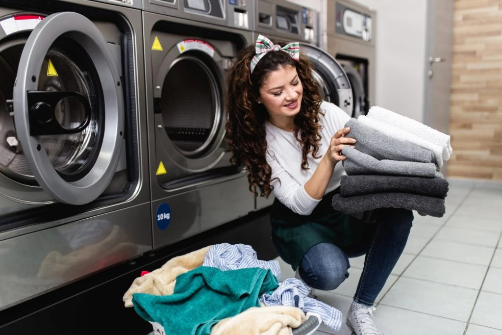 A woman kneels in front of washing machines, holding a stack of folded towels, with laundry on the floor, as she dreams of the convenience that laundry pick-up services could bring to her hectic life.