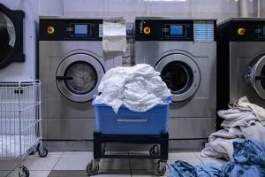 A laundry room with two industrial-sized washing machines; a blue bin filled with white linens and piles of laundry are visible on the floor, showcasing the efficiency of our Wash and Fold Laundry Service.