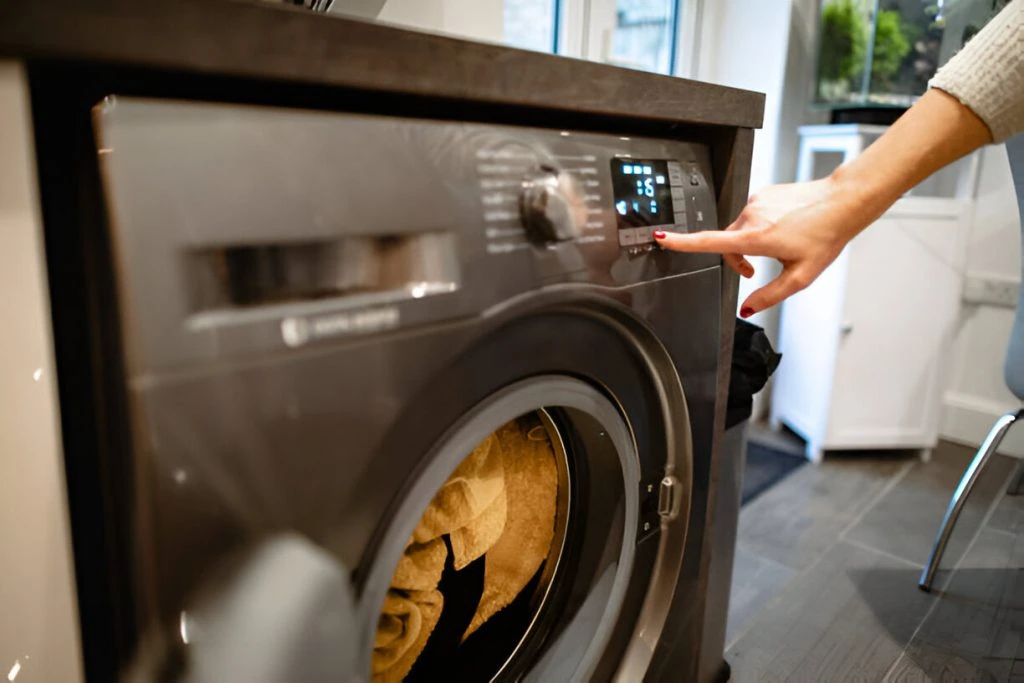 A person adjusts the settings on a front-loading washing machine with towels inside, preparing them for dry cleaning pick-up.