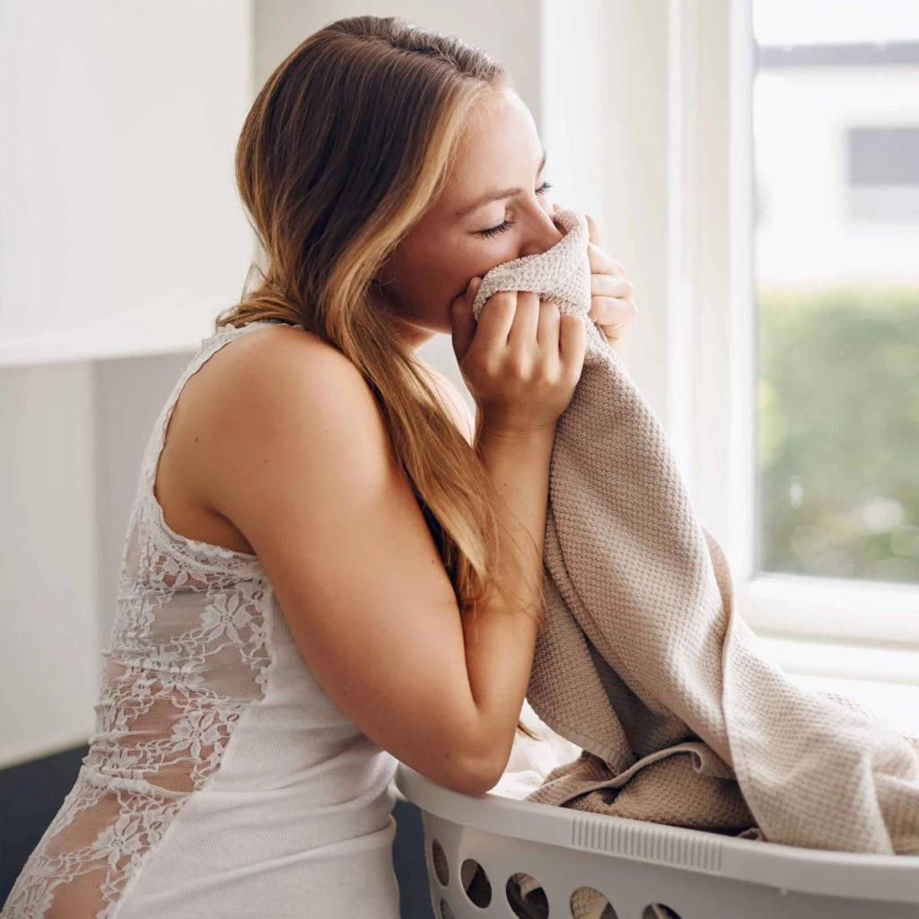 Woman in a lace dress smells a towel while leaning on a laundry basket near a window, savoring the freshness right before her dry cleaning pick-up.