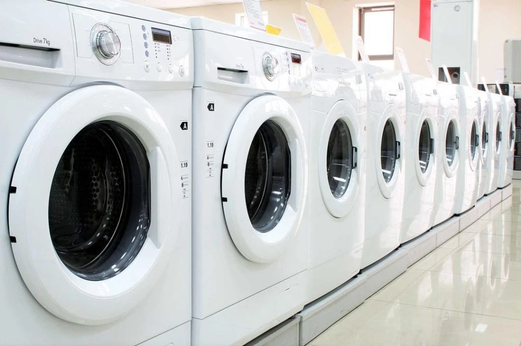 A row of white front-loading washing machines is displayed in a store, perfectly aligned on the shiny floor, offering seamless efficiency alongside dry cleaning delivery services.