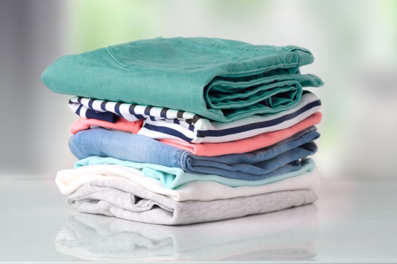 A neatly folded stack of clothes, including a green shirt, striped tops, and various colored garments, awaiting pick-up for the dry cleaning delivery service on a pristine white surface.