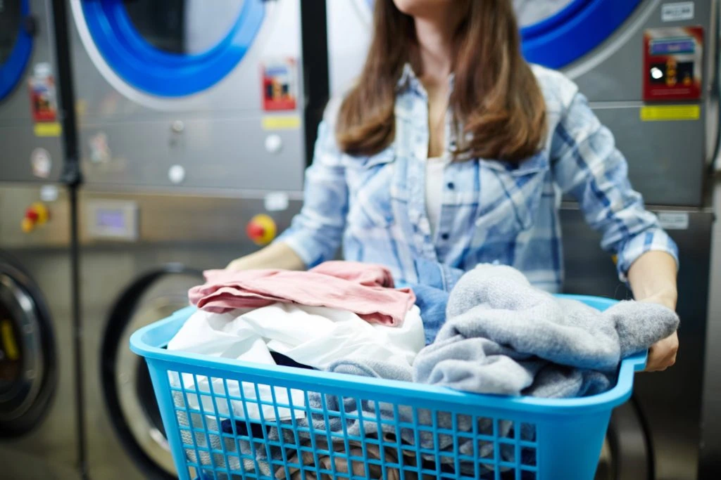 A person in a plaid shirt holds a blue laundry basket brimming with clothes, contemplating the convenience of wash and fold services at the local laundromat.