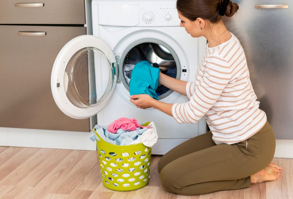 A woman kneels in front of a washing machine, placing clothes inside with meticulous care. Beside her on the wooden floor rests a green laundry basket, exemplifying perfect laundry organization as it holds a neatly arranged collection of garments.