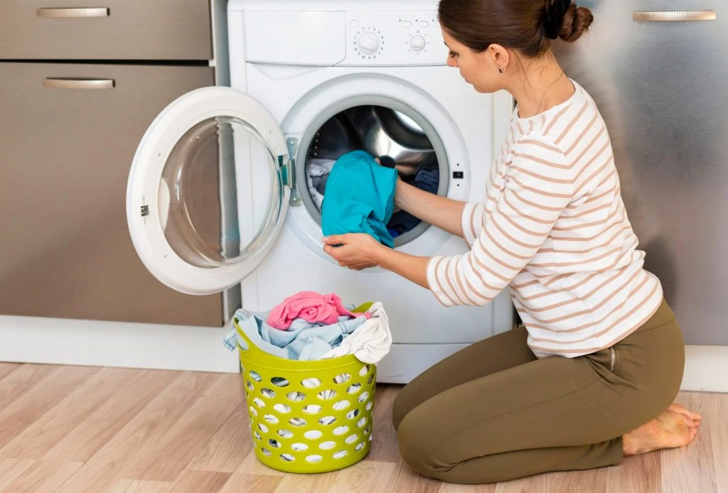A woman kneels in front of a washing machine, placing clothes inside with meticulous care. Beside her on the wooden floor rests a green laundry basket, exemplifying perfect laundry organization as it holds a neatly arranged collection of garments.