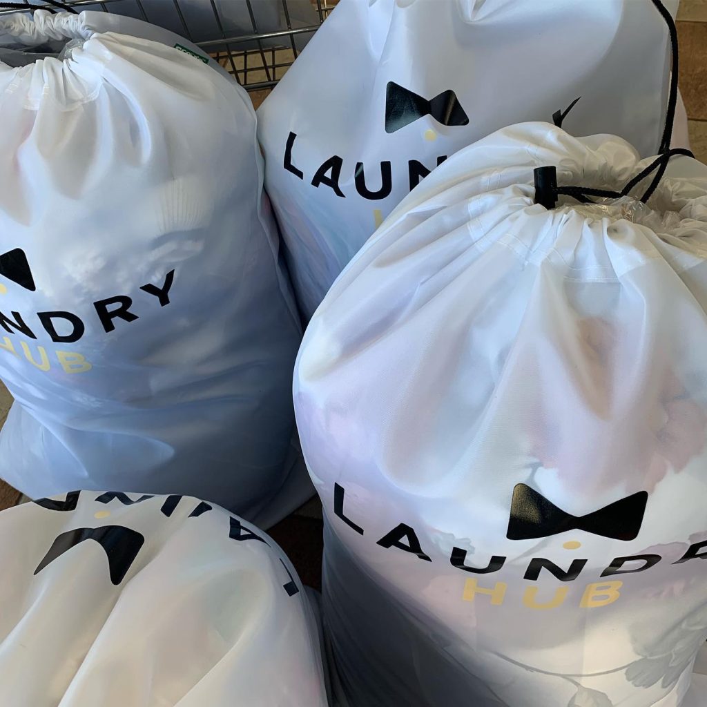 Four white laundry bags with "Laundry Hub" written on them, expertly filled and cinched at the top, showcase impeccable laundry organization as they rest neatly on a surface.