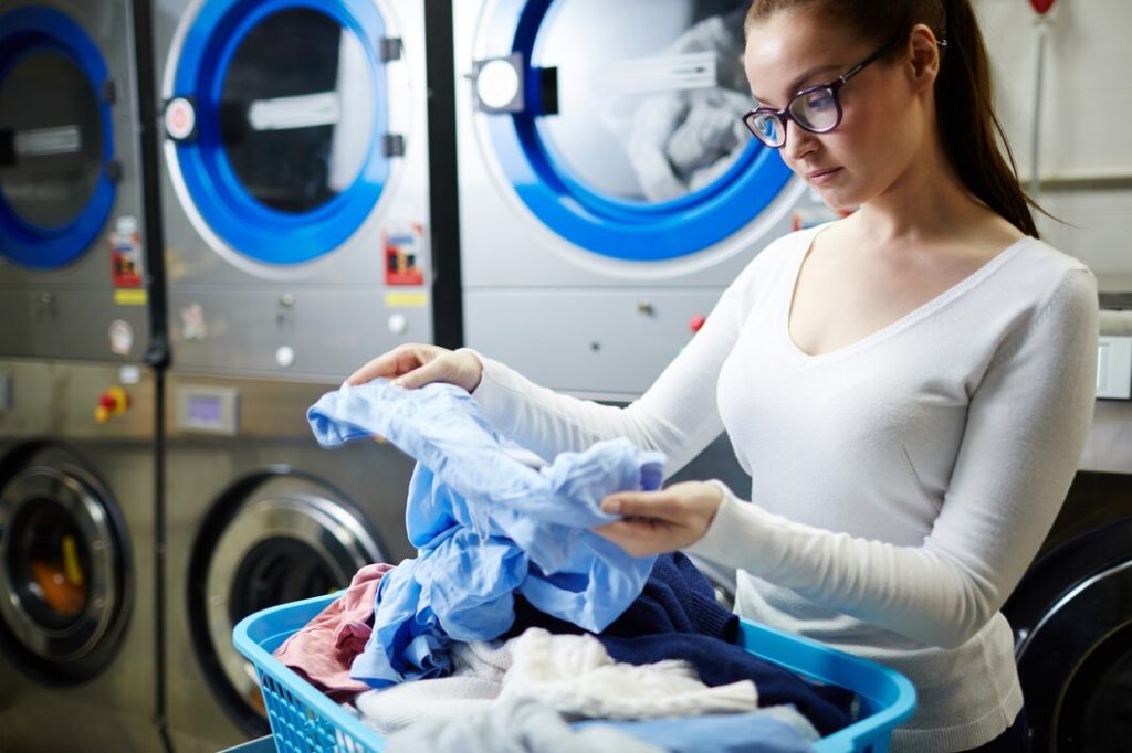 A woman stands in a laundromat, holding laundry in front of a whirring washing machine.